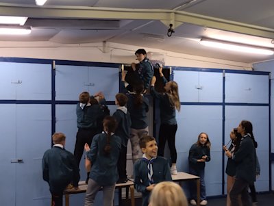 A group of Scouts have made themselves into a tower, standing on a table and are helping one of the Scouts, who is a bit of a climbing monkey, to stay on the built-in cupboard and hold the team's potato in place. They were set a task to make the highest tower using a potato. The remaining Scouts are watching and several Scouts on the other team seem to find this all very funny.