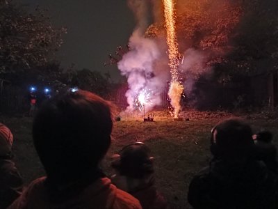 Two Beavers with ear defenders on and a Scout watch the fireworks display. The shot cake has left a streak of sparks as it sent a shot into the air, while the Catherine wheel spins brightly beside it.