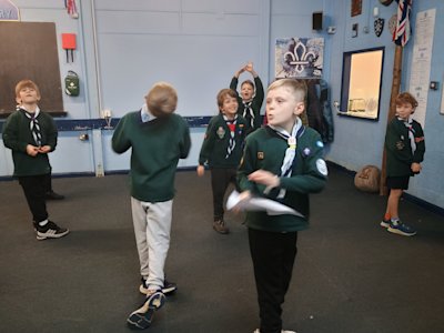 The Cubs are playing a version of Granny Footsteps where they have to spell their names to move forward, as a leader calls out the phonetic alphabet letters randomly. Several Cubs seem excited by the latest letter called.