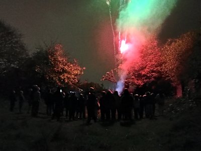 The Scout group is watching the fireworks display together. The colours of the shot cake are giving an eerie glow to the trees behind them.