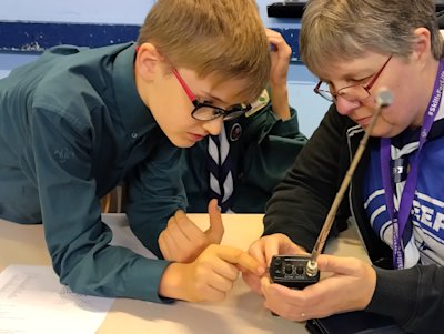 A Scout looks intently at the walkie talkie the leader is holding, reaching out to press the button.