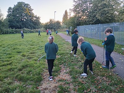 A group of Cubs are spread out along the grass and path, litter pickers in hand, looking for any scraps of rubbish they can find. The sun is low in the sky and the Cubs have smiles on their faces.