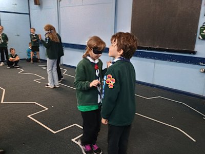 One Cub is blindfolded and making her way along a taped zigzag line, guided by the voice and instructions of her fellow Cub who has a cheeky grin on his face. In the background, we can see another pair of Cubs finishing their run of the taped line while others wait their turn.