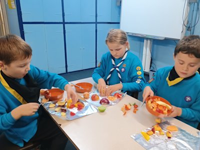 Three Beavers are sat at a table, chopping up a variety of fruit. One Beaver is looking excited about preparing his nectarine, another tips his bowl of fruit onto a piece of foil, while the third begins pulling her orange into segments.