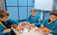Three Beavers are sat at a table, chopping up a variety of fruit. One Beaver is looking excited about preparing his nectarine, another tips his bowl of fruit onto a piece of foil, while the third begins pulling her orange into segments.