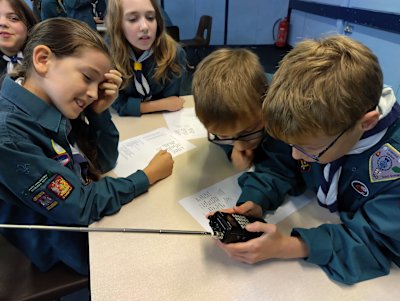 A group of Scouts cluster around a walkie talkie. One boy holds it in both hands while another watches at his elbow. Three girls watch the boys, with various expressions of interest and amusement on their faces.