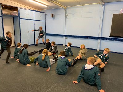 Two rows of Scouts are seated spaced on the floor, legs straight out in front of them, feet touching the feet of the Scout opposite them. Two Scouts are running the length of the rows, racing each other.