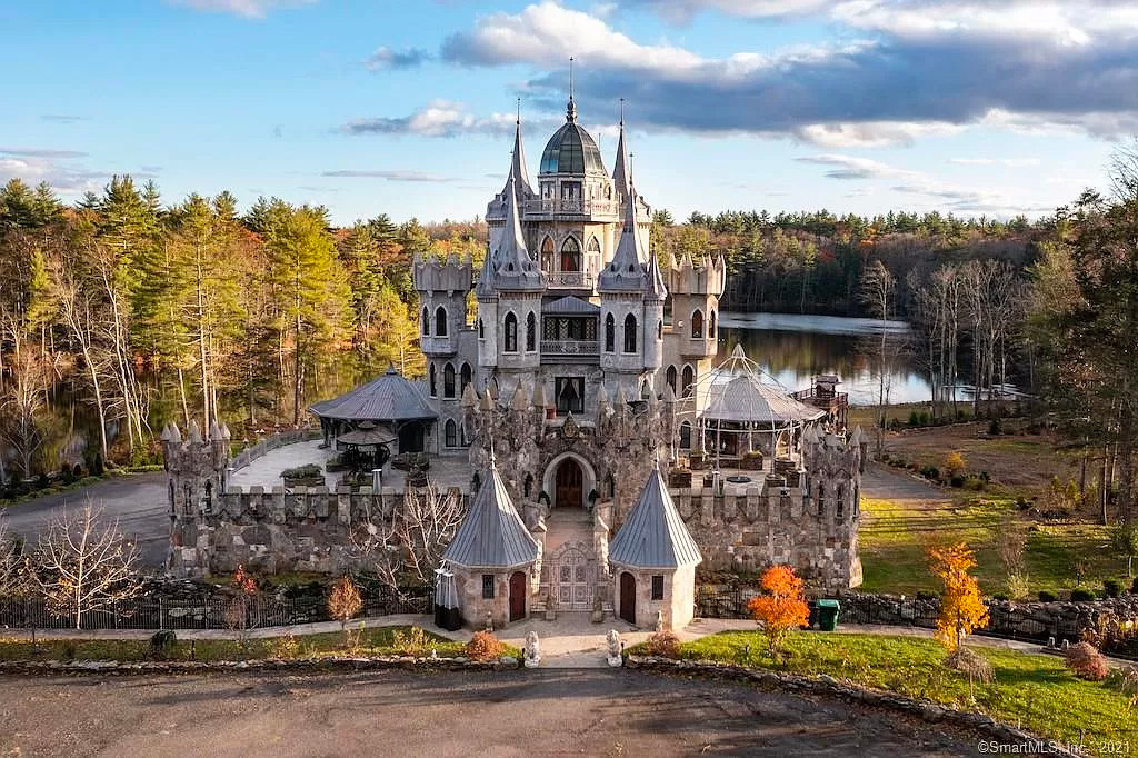 Front view of a home that looks like a castle in front of a lake and surrounded by trees.