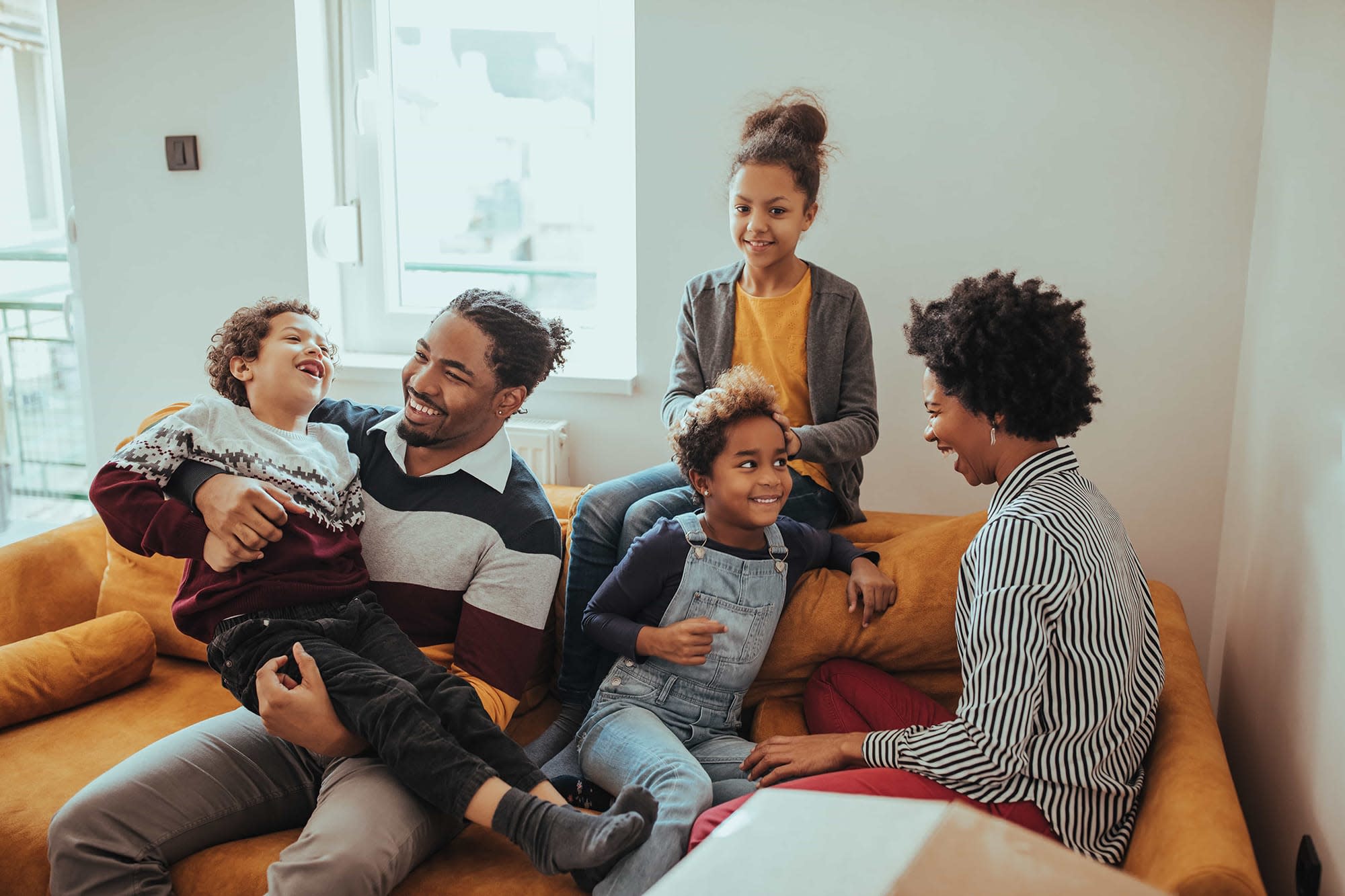 Happy family of five laughing and spending time together on a yellow couch in a bright, cozy living room.
