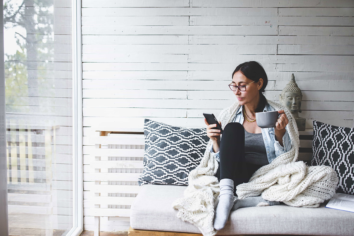 Woman sitting on couch wrapped in a blanket looking at her phone in one hand and holding a coffee mug in the other hand. 