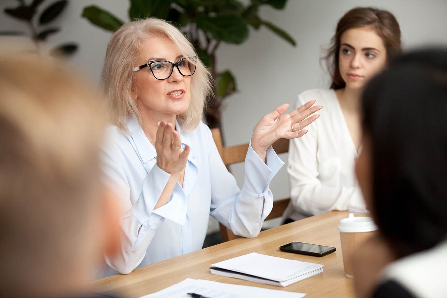 Older woman with a blue long-sleeved shirt and glasses talking to a table full of people.