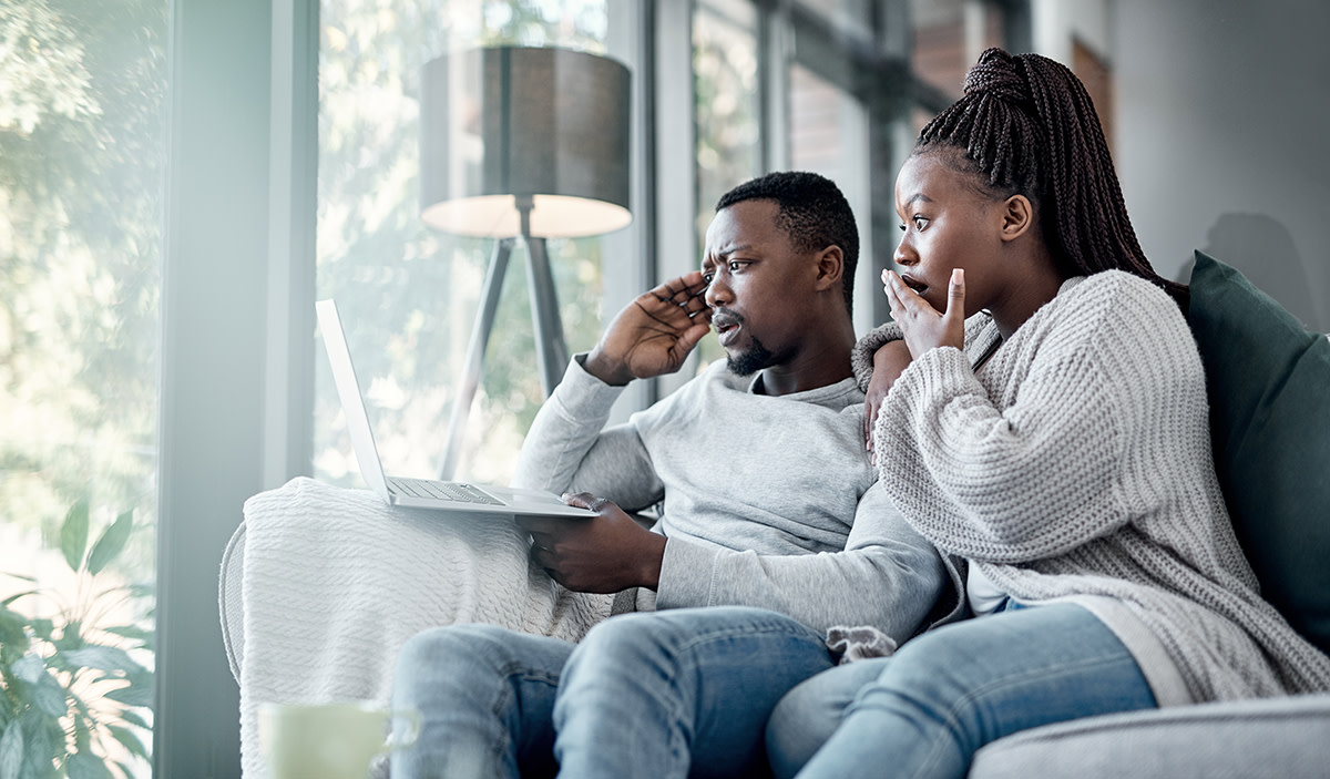 Man and woman sitting on a couch and looking at a laptop with shocked expressions.