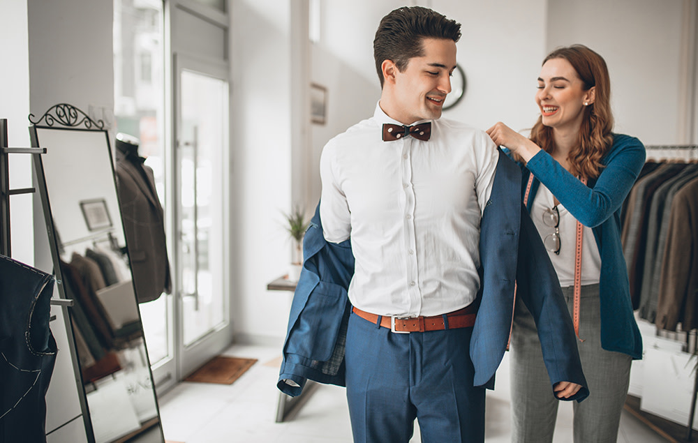 Store associate assists man in putting on suit jacket in suit store.