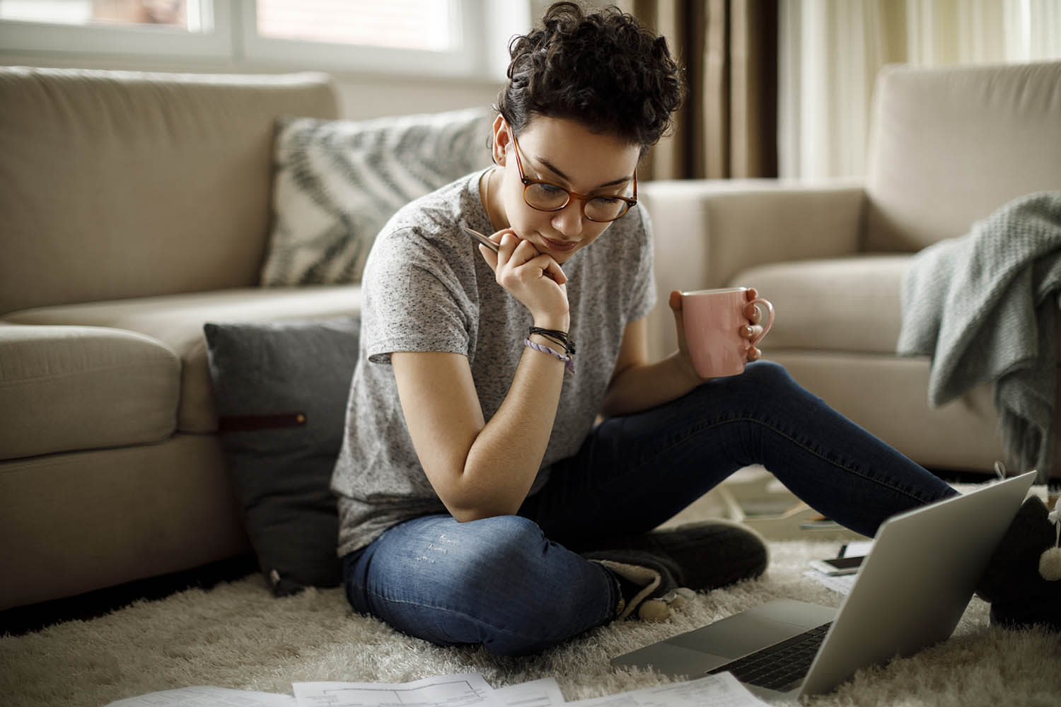 A woman sitting on her floor in front of a computer focused on papers below her. 
