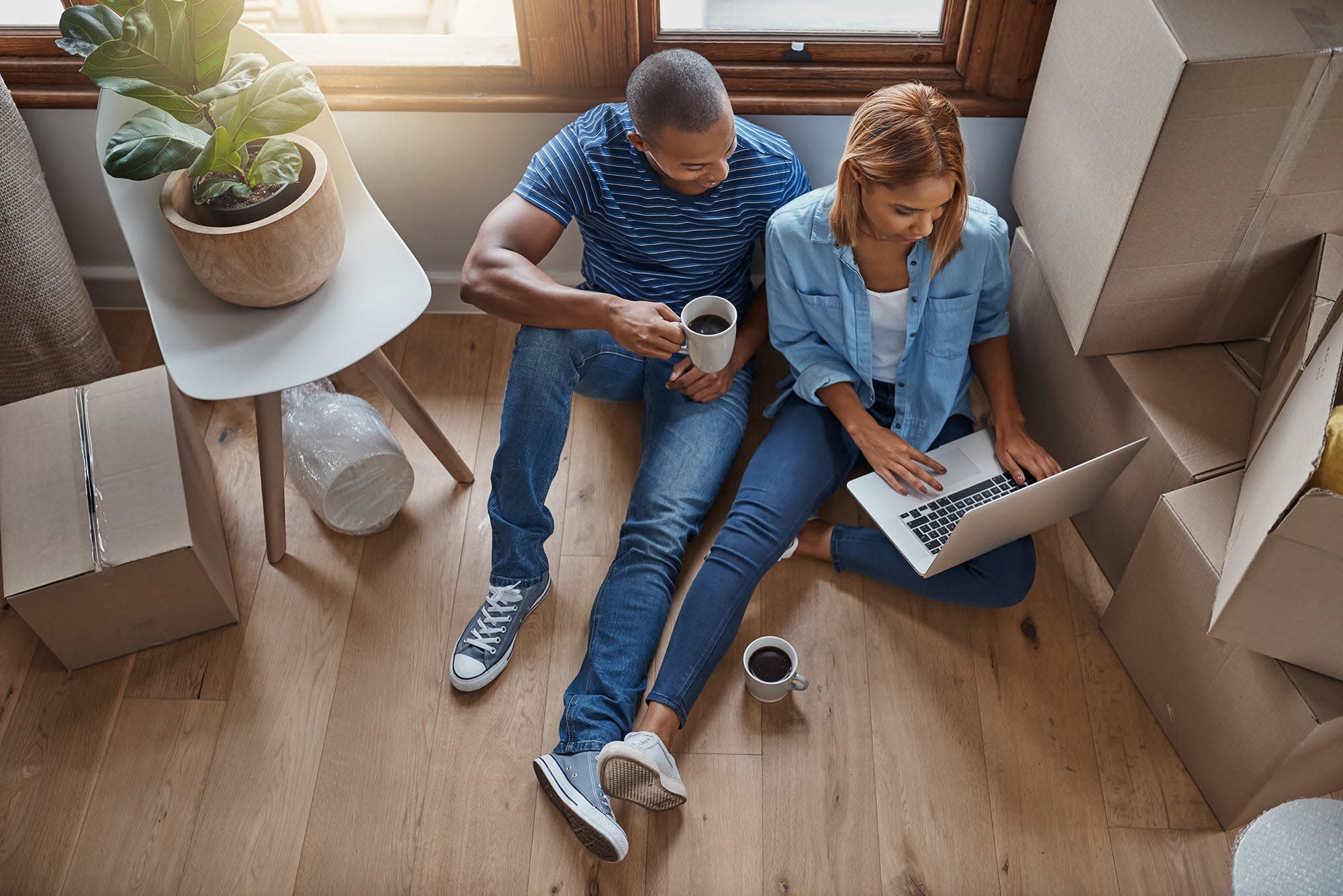 A couple sitting on the floor of their home surrounded by moving boxes browsing their computer. 