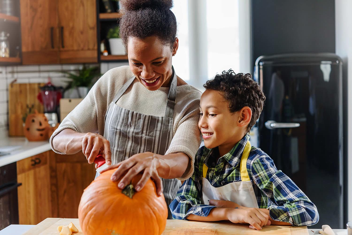 Mother and son wearing aprons while mother carves a pumpkin and son watches. 