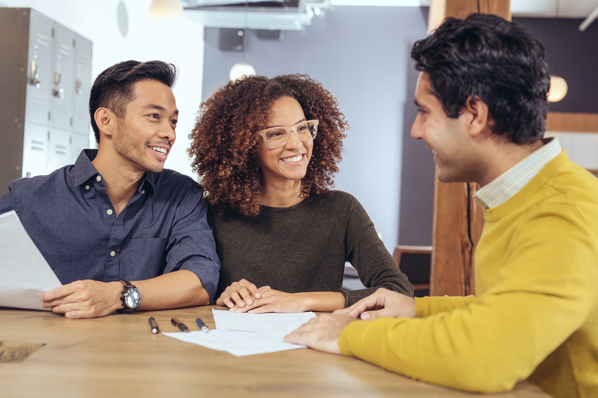 Smiling couple meets with a real estate agent at a table to review and discuss documents in a modern office setting.