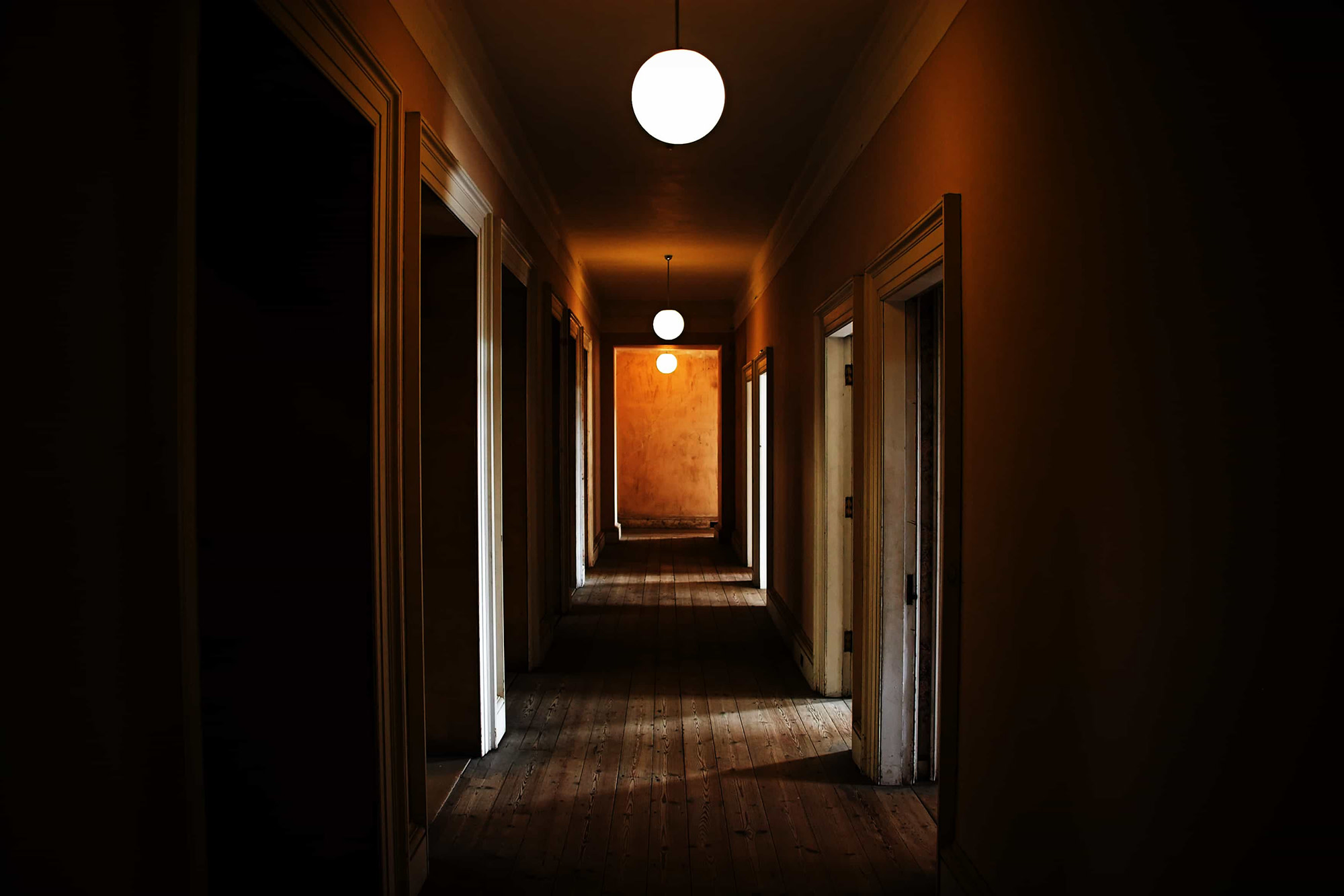 A dark hallway dimmed by three circular lights hanging from the ceiling and doors to rooms on each side of the hallway.