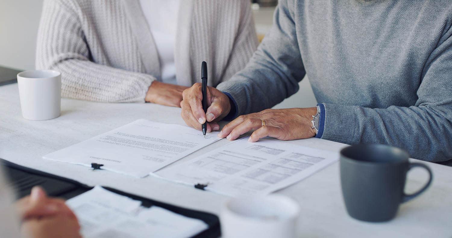 Two people sitting next to each other at a table while one signs a stack of papers.
