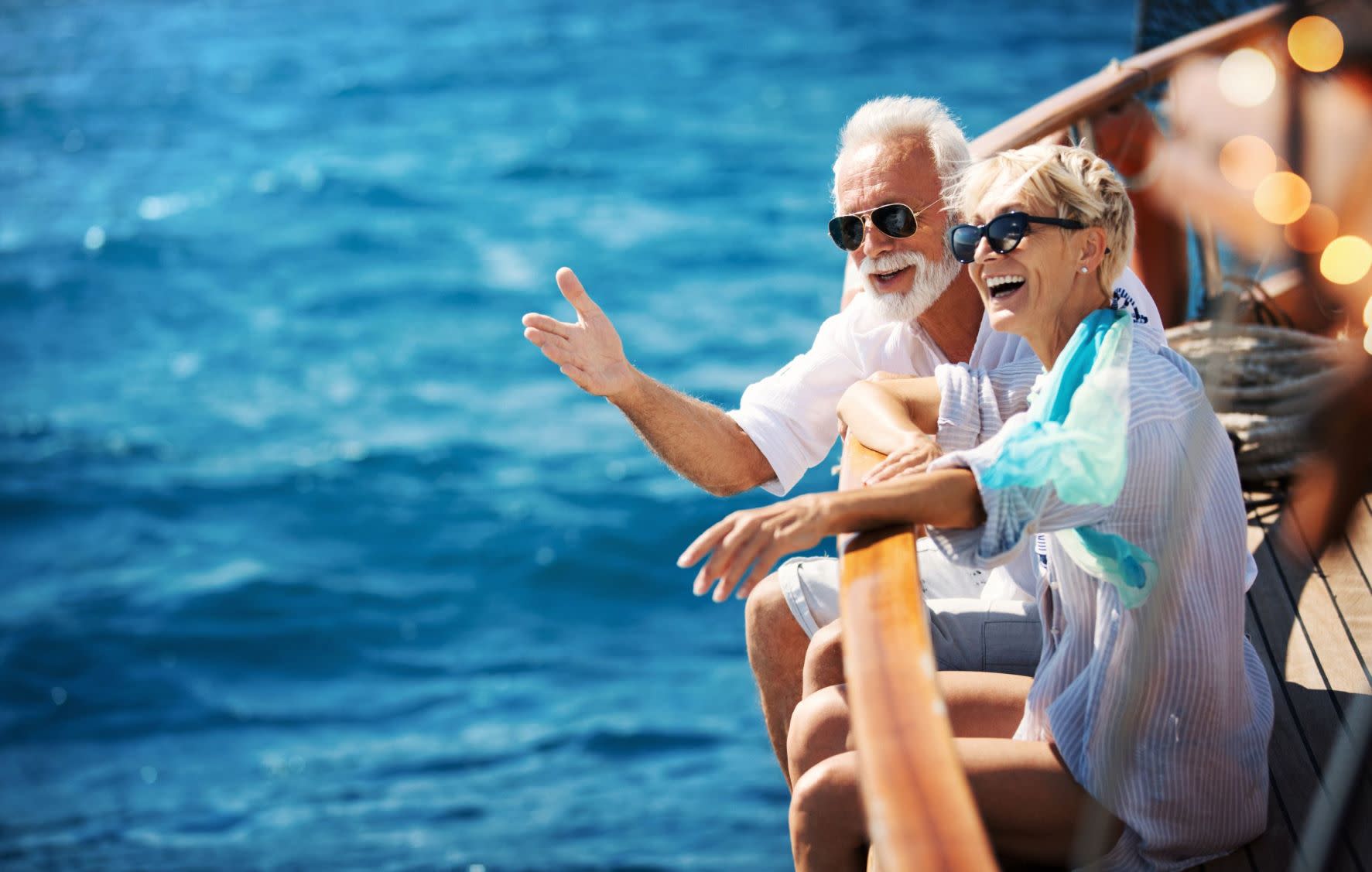 Older couple wearing sunglasses sitting on floor of boat with their legs dangling over the side and arms resting on railing. 