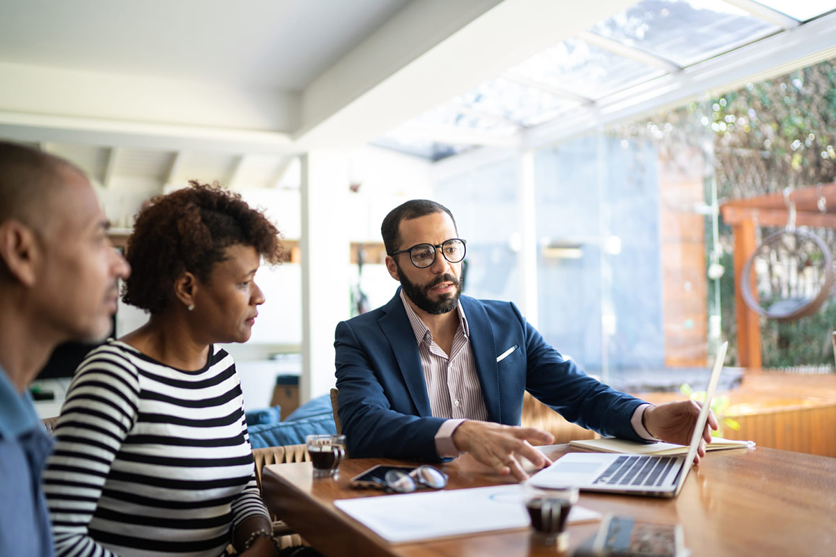 Three people sitting at a desk in front of an open laptop while one of them explains what is on the laptop.