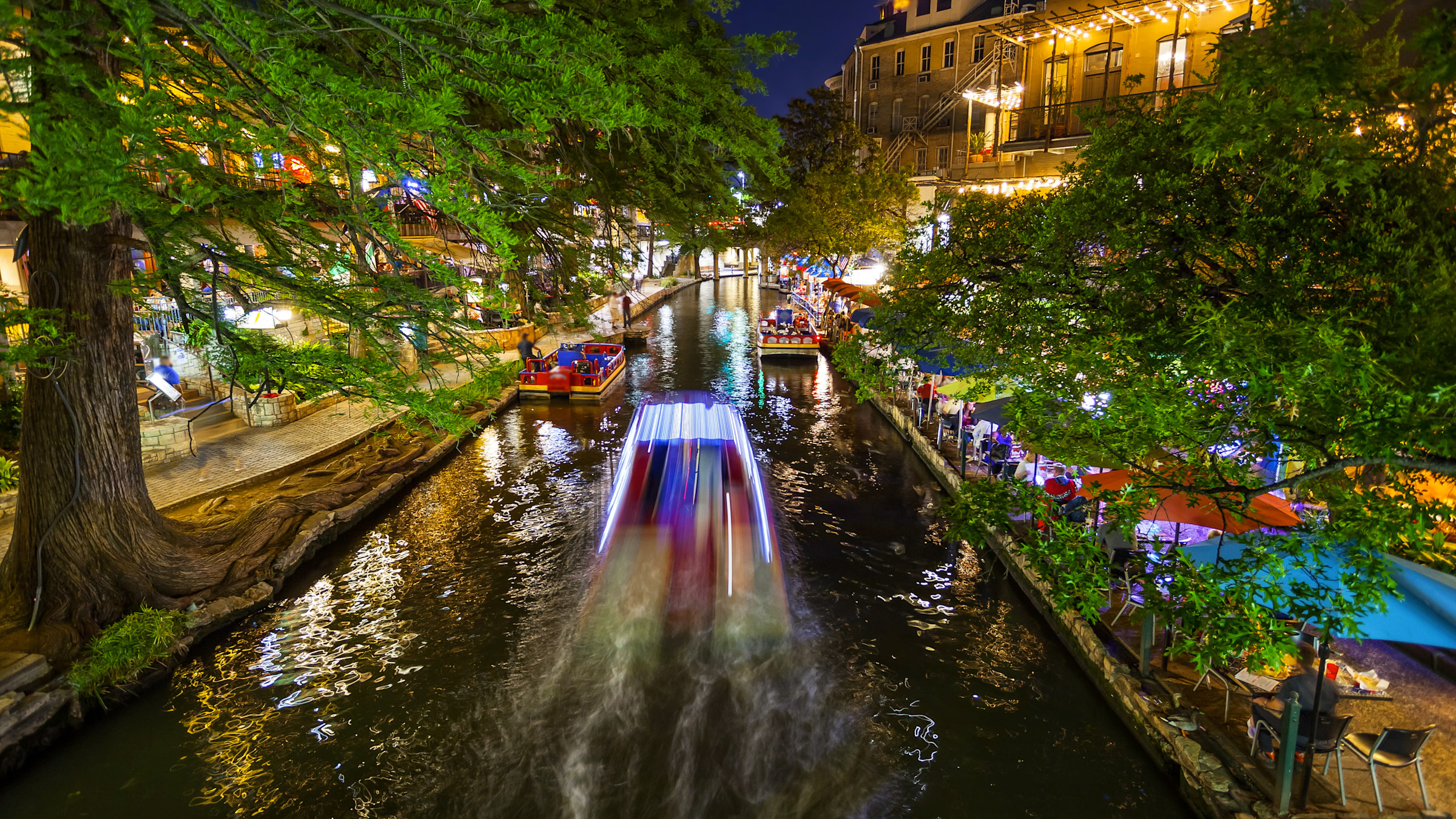 Night image of a river passing through city with buildings on each side and small boats on the river.