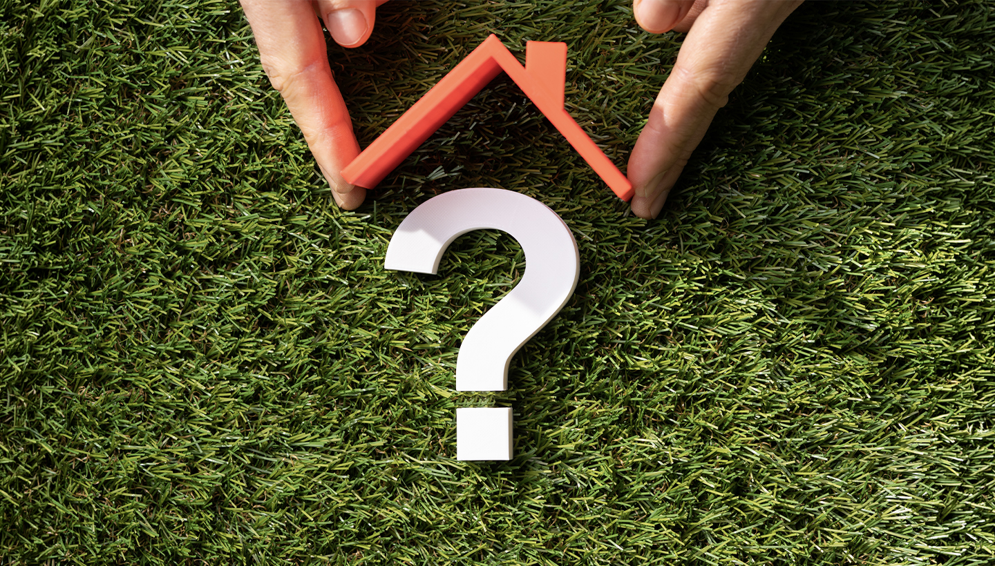 Hands holding a red house roof shape over a white question mark on green grass, symbolizing uncertainty in homeownership or real estate decisions.