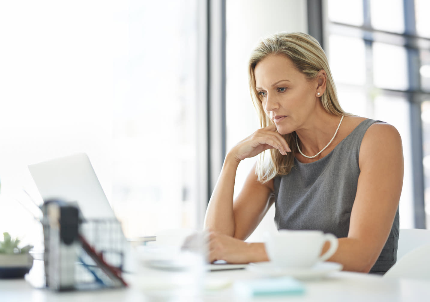 Woman sitting at table with hand on chin looking at open laptop.