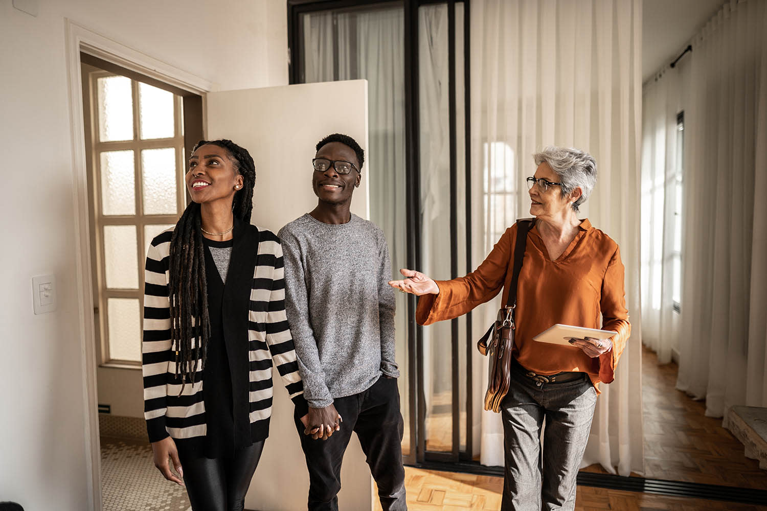 Couple holding hands looking around the inside of a house while an older woman holding a tablet explains.