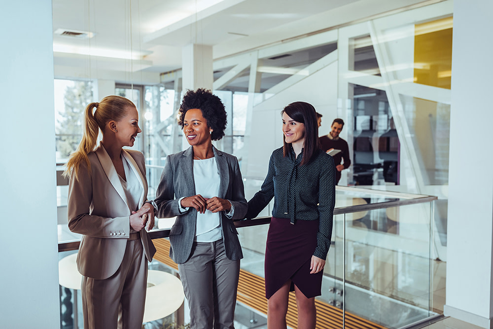 Three women dressed in professional attire in an office talking to each other.
