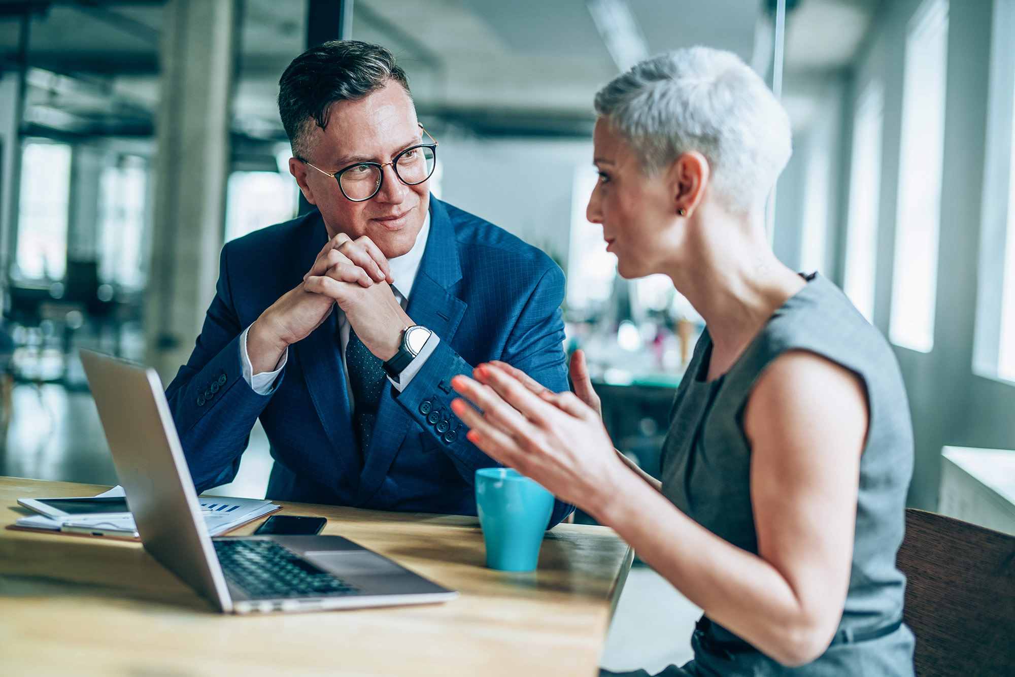 Man and woman sitting at table in office meeting room having a conversation in front of an open laptop.