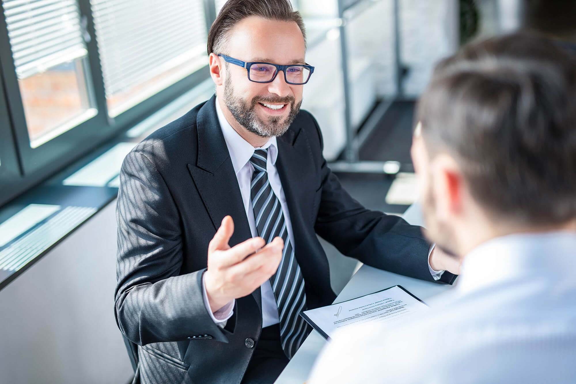 Smiling man wearing glasses and a suit and tie talking to another man facing him.