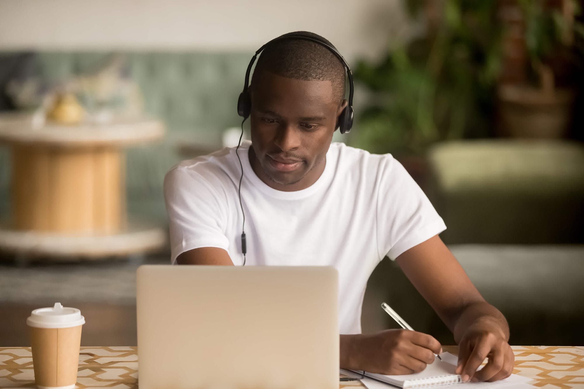 A man with headphones on looking at the computer screen taking notes on a note pad. 