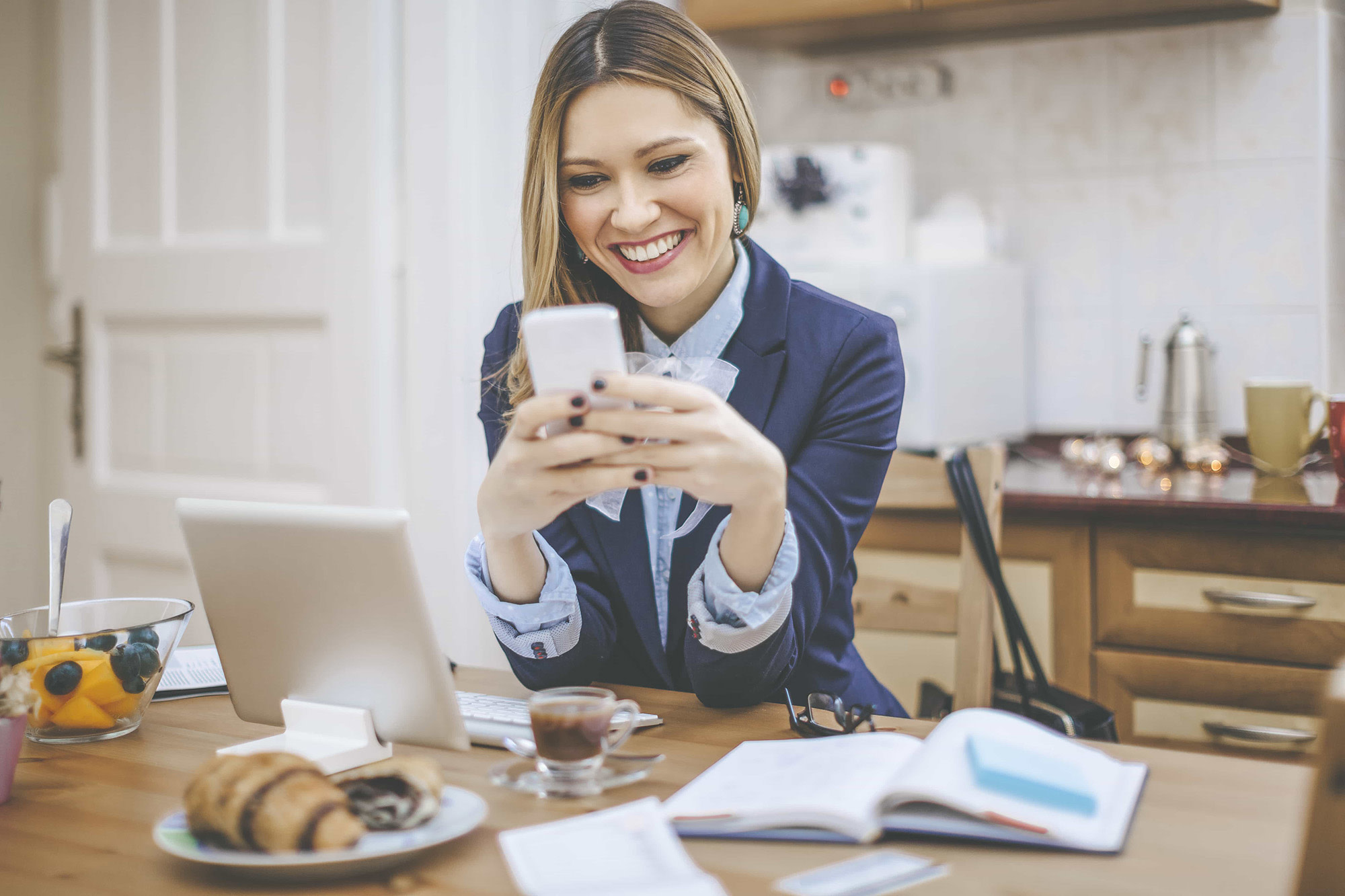 Woman sitting at kitchen table in front of a propped up tablet and smiling while looking at phone in hands. 