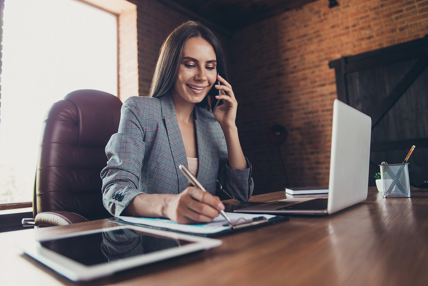 Woman in office sitting at a desk with an open laptop is talking on the phone and writing on a sheet of paper attached to a clipboard.