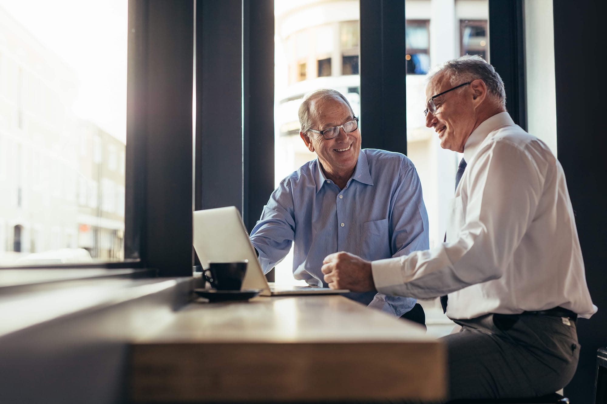 Two older men wearing glasses and business professional attire talking to each other while sitting at a window facing table in front of a laptop.