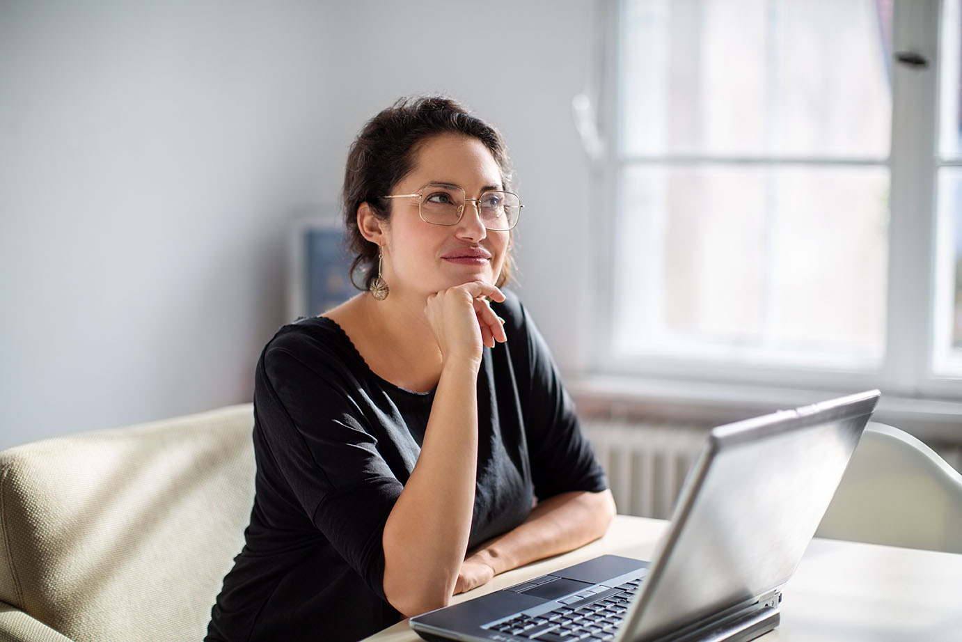 Woman sitting at desk in front of an open laptop with a hand to chin and a thinking expression on her face.