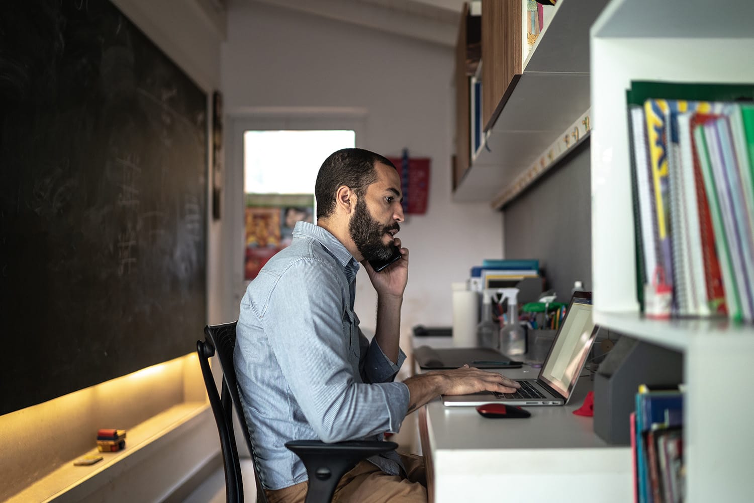 A man on his phone and his computer sitting at his desk at home. 