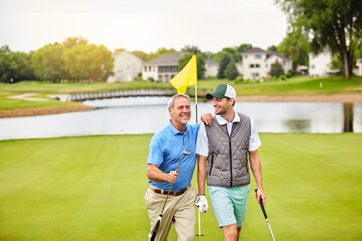 Two men on a golf course holding golf clubs are walking away from a golf putting green flag.