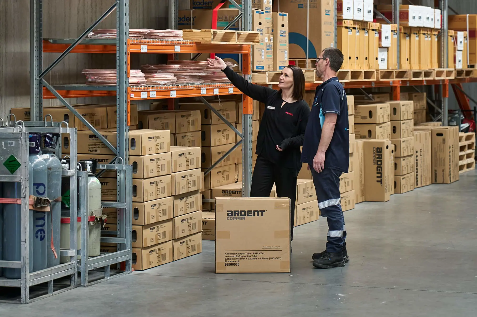 Woman in black uniform pointing at shelves while standing with man in blue uniform in warehouse with stacked boxes.