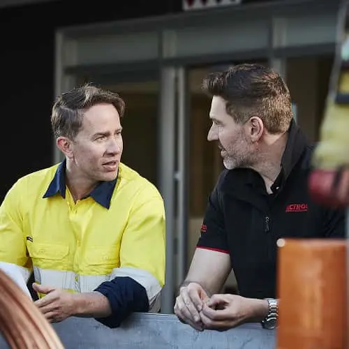 Two men in work attire having a conversation, one wearing a yellow safety shirt and the other in black.