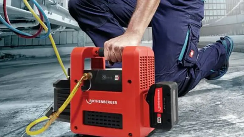 Technician in blue uniform kneeling beside red Rothenberger vacuum pump with yellow hose on industrial floor