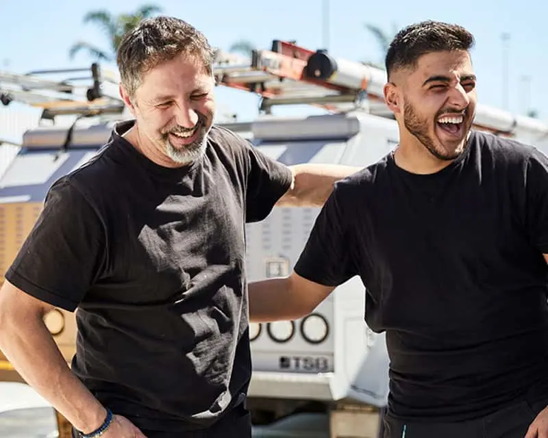 Two men in black t-shirts laughing together outdoors with a work truck and palm trees in the background.