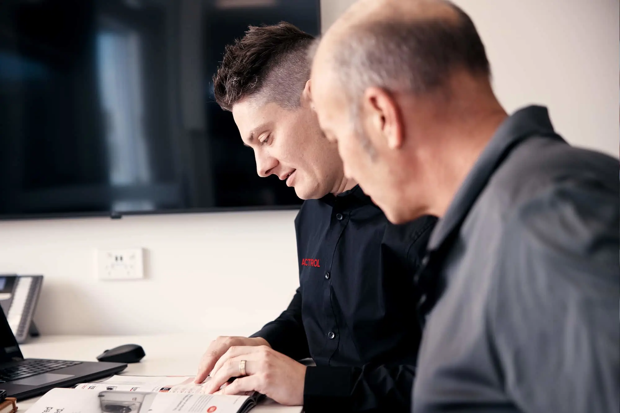 Two men in dark shirts reviewing documents at a desk with a laptop and computer monitor in the background.
