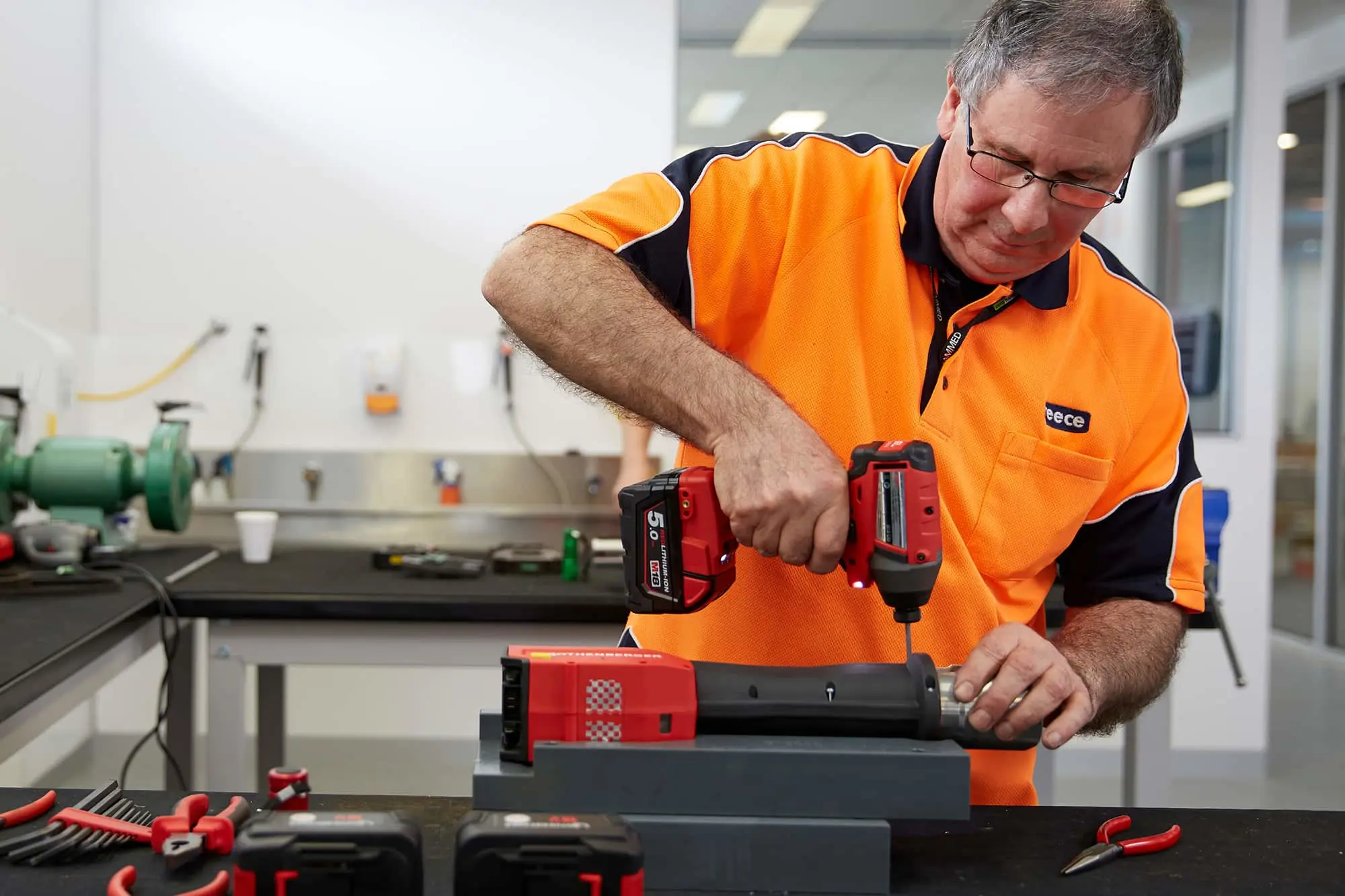 Worker in orange safety shirt using a red power drill on equipment in a workshop with tools on the workbench.