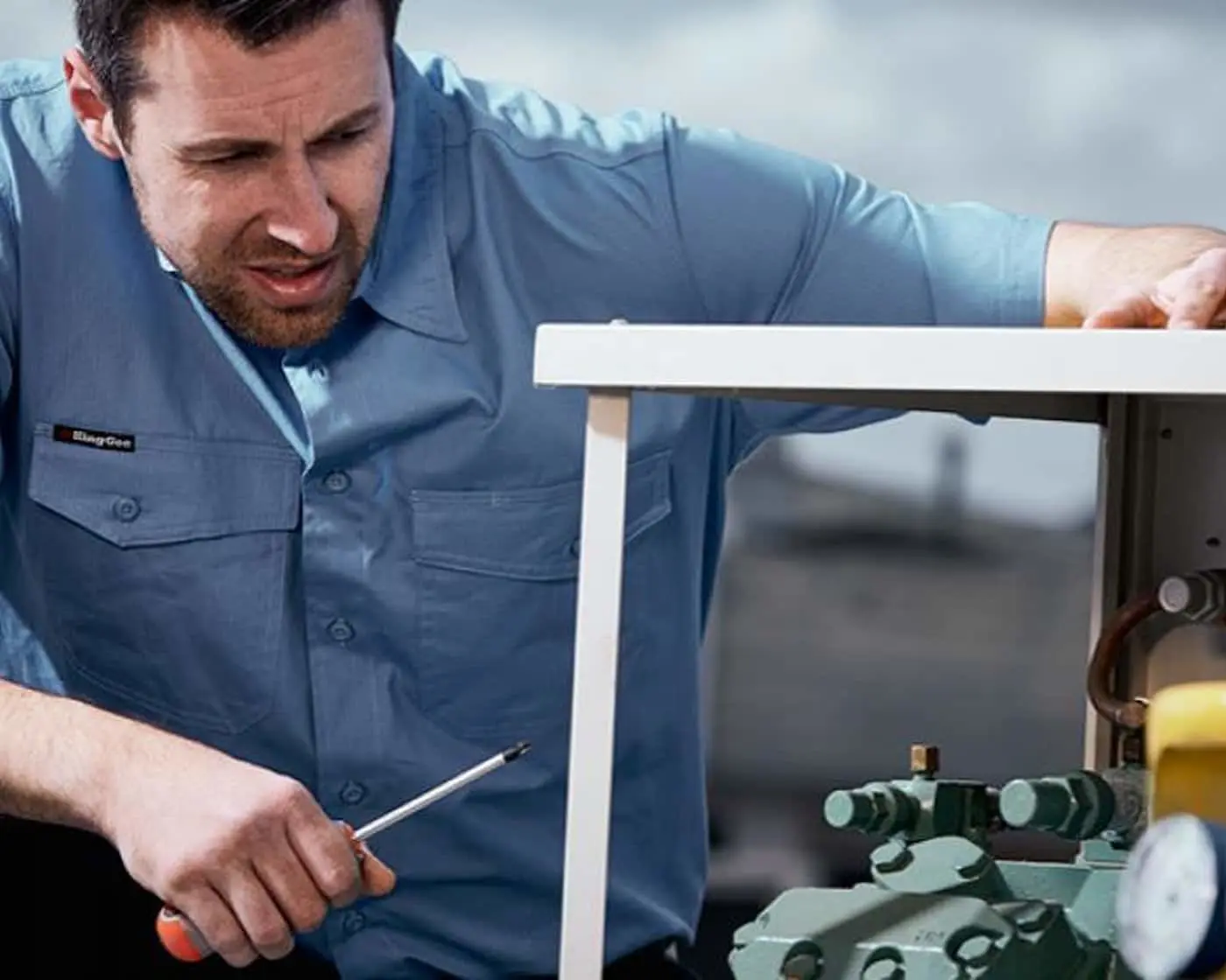 Man in blue work shirt using a tool to repair or service equipment with green mechanical components.