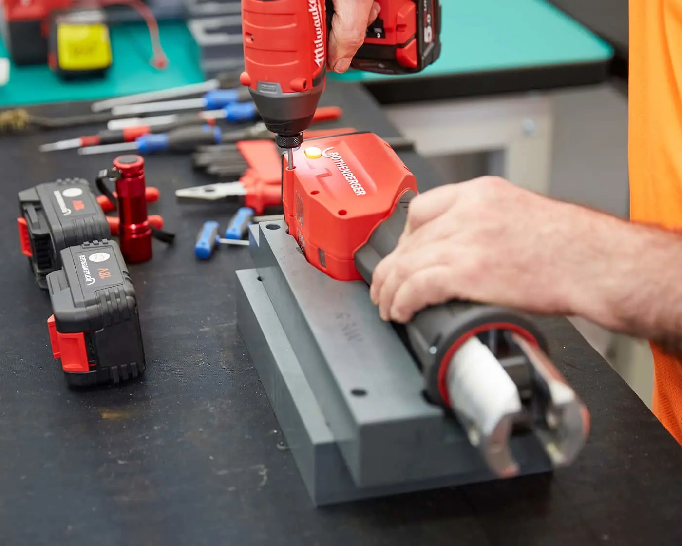 Person using red cordless drill on black workbench with various tools and equipment scattered around.