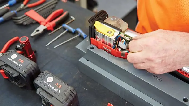 Person in orange shirt repairing a power tool battery on a workbench with various tools and batteries nearby.