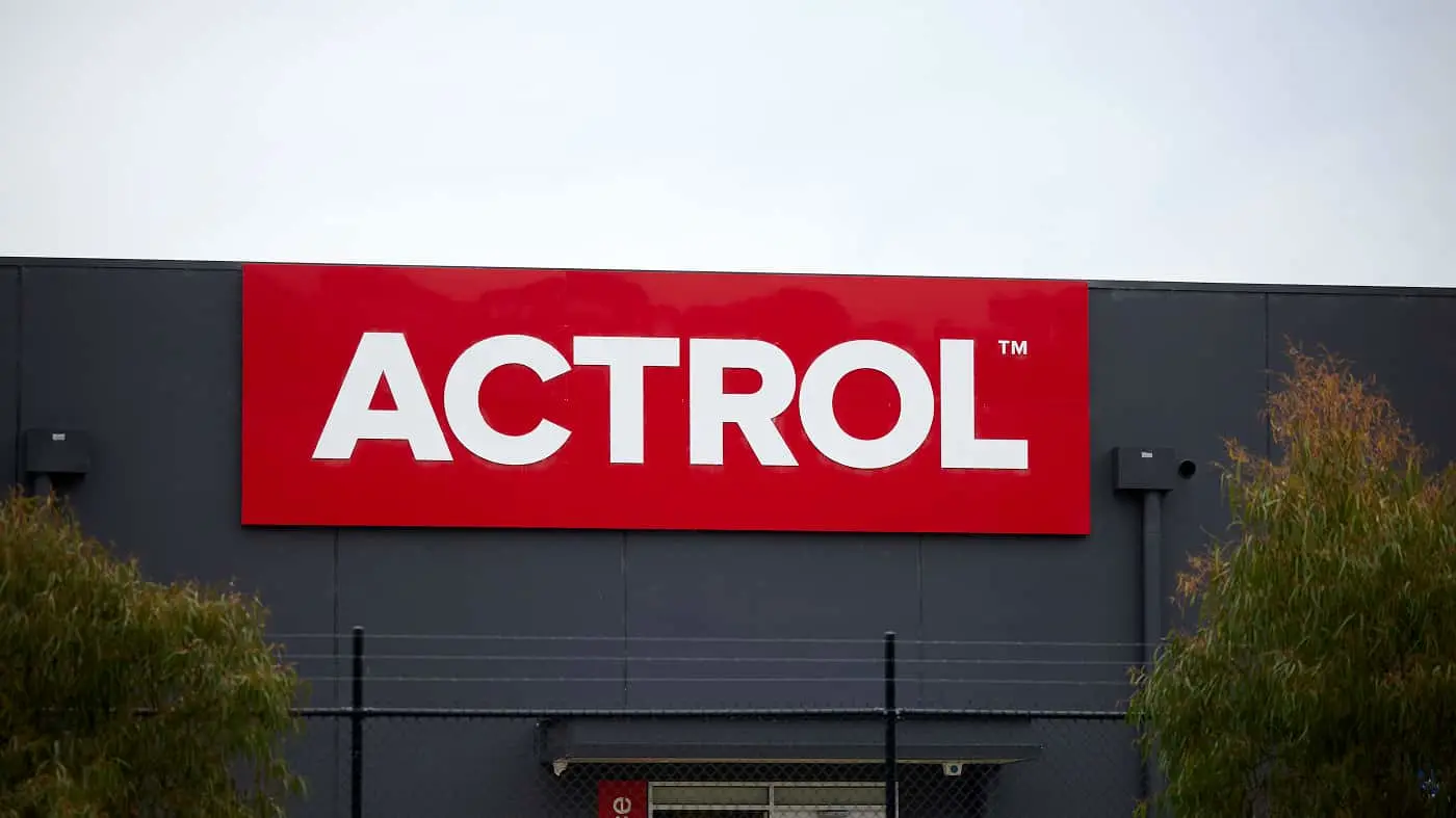 Red ACTROL sign with white letters mounted on a dark gray building exterior with plants on either side.