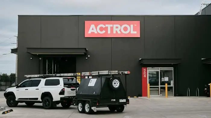 White pickup truck with roof rack and trailer parked in front of dark gray ACTROL building with red signage